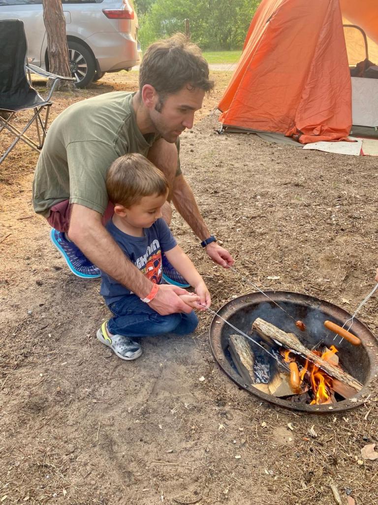 Father and son cooking hot dogs on a campfire.